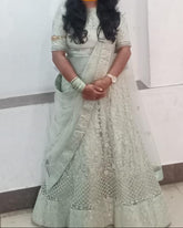 A woman wearing a light sage green embroidered traditional outfit with a matching dupatta, standing indoors against a plain white wall. Her face is obscured by a yellow smiling emoji, and a brass oil lamp is visible on the right side.