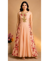 A woman modeling a peach-colored sleeveless Anarkali gown with green and red floral embroidery on the bodice and flared skirt, set against a warm beige background.