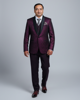 A man modeling a dark purple brocade suit with black satin lapels, a matching vest, a patterned tie, and dark brown dress shoes, photographed against a solid gray studio background.