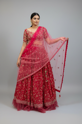A woman modeling a traditional Indian red lehenga choli set with a sheer pink dupatta, photographed against a solid gray studio background.
