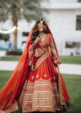Woman in a traditional red-and-gold Sabyasachi bridal lehenga with a veil, outdoors.