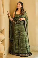 A woman (Sara Tendulkar) models a green printed traditional outfit with gold embroidery from Anita Dongre, consisting of a sleeveless top, flared pants, and a matching draped dupatta, set against a beige architectural background.