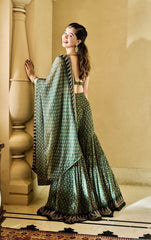 A woman (Sara Tendulkar) wearing a green patterned lehenga choli saree from Anita Dogre stands in a warm-toned indoor setting, posing next to a decorative pillar.