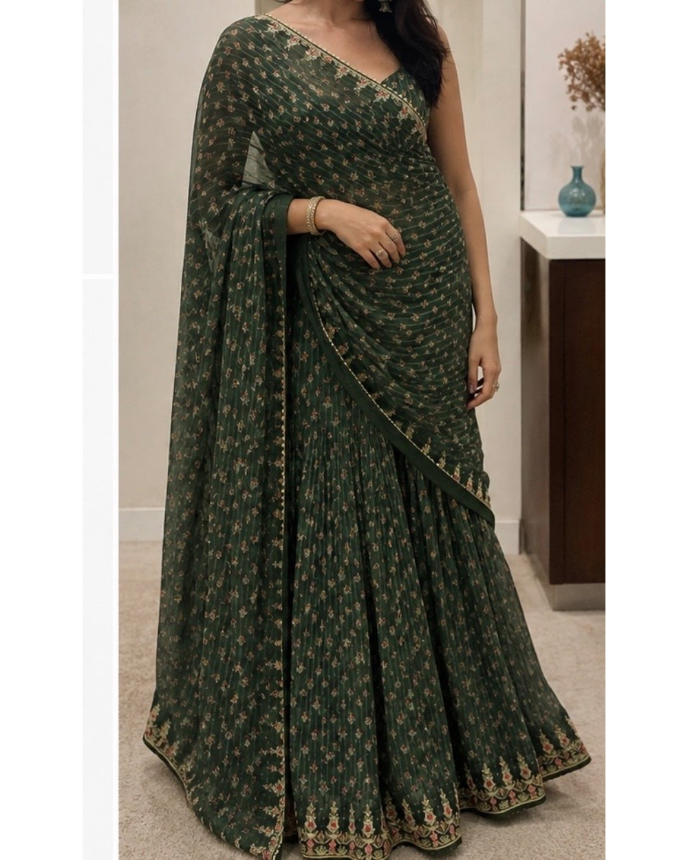 A woman wearing a dark green georgette lehenga choli saree with gold floral prints and border embroidery, standing in an indoor setting.