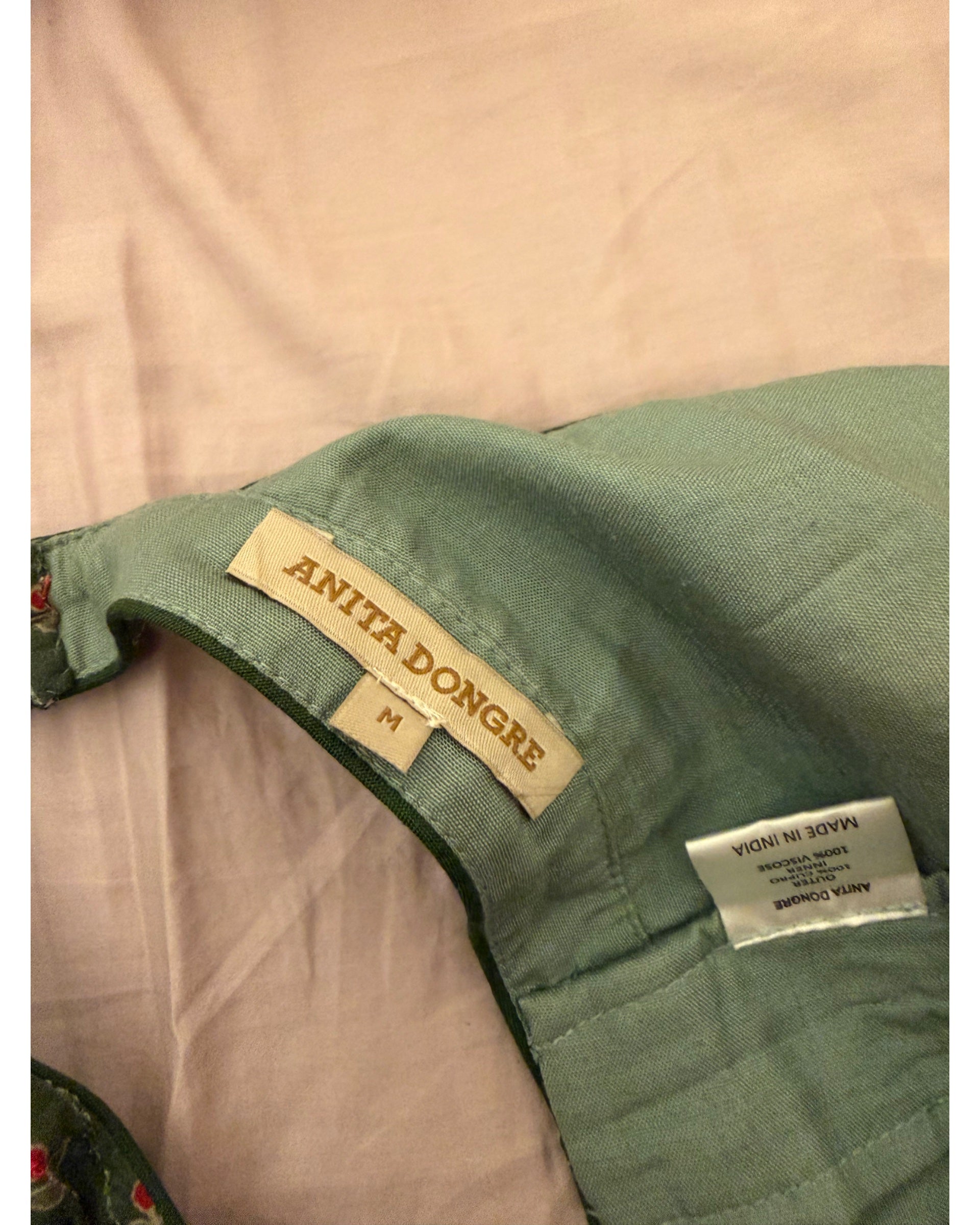 Close-up of a green quilted garment with visible Anita Dongre brand labels and a size M tag, laid on a light pink fabric background.