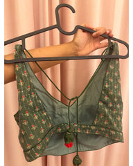 Back view of a dark green printed blouse top with red and gold floral motifs, held on a black hanger by a person's hand against a pink curtain backdrop.