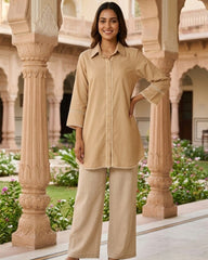 A woman modeling a beige long-sleeve kurta with matching wide-leg trousers, standing in a traditional courtyard with stone pillars and greenery.