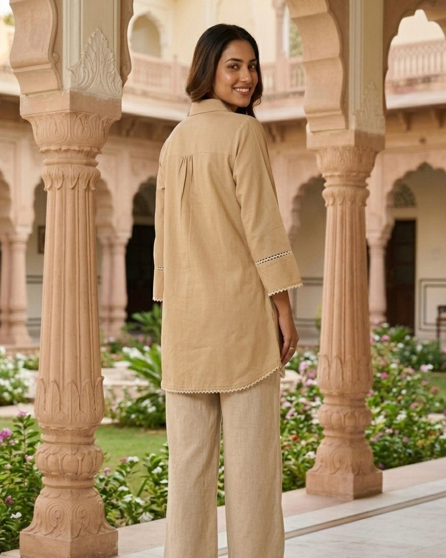 A woman wearing a beige kurta and matching wide-leg trousers stands in a courtyard with stone pillars and greenery in the background.