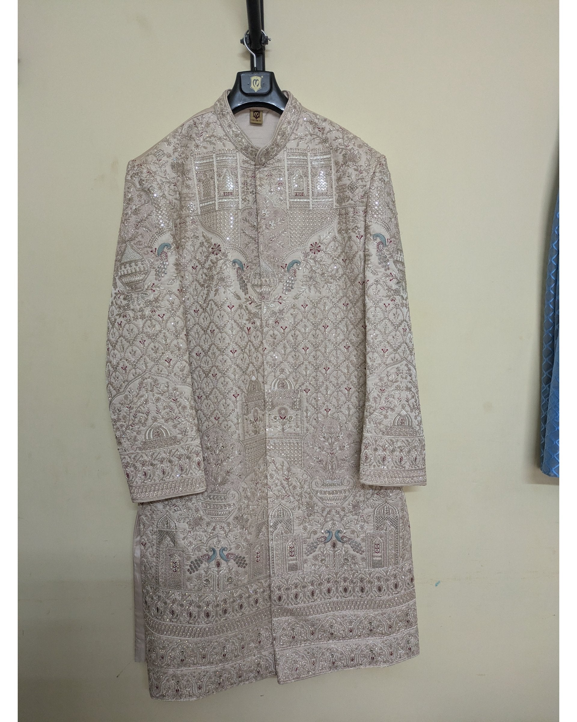 A beige embroidered sherwani jacket with intricate geometric patterns and small blue floral accents, hanging on a black hanger against a plain beige wall.