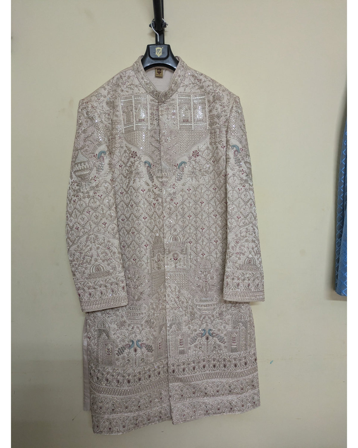 A beige embroidered sherwani jacket with intricate geometric patterns and small blue floral accents, hanging on a black hanger against a plain beige wall.