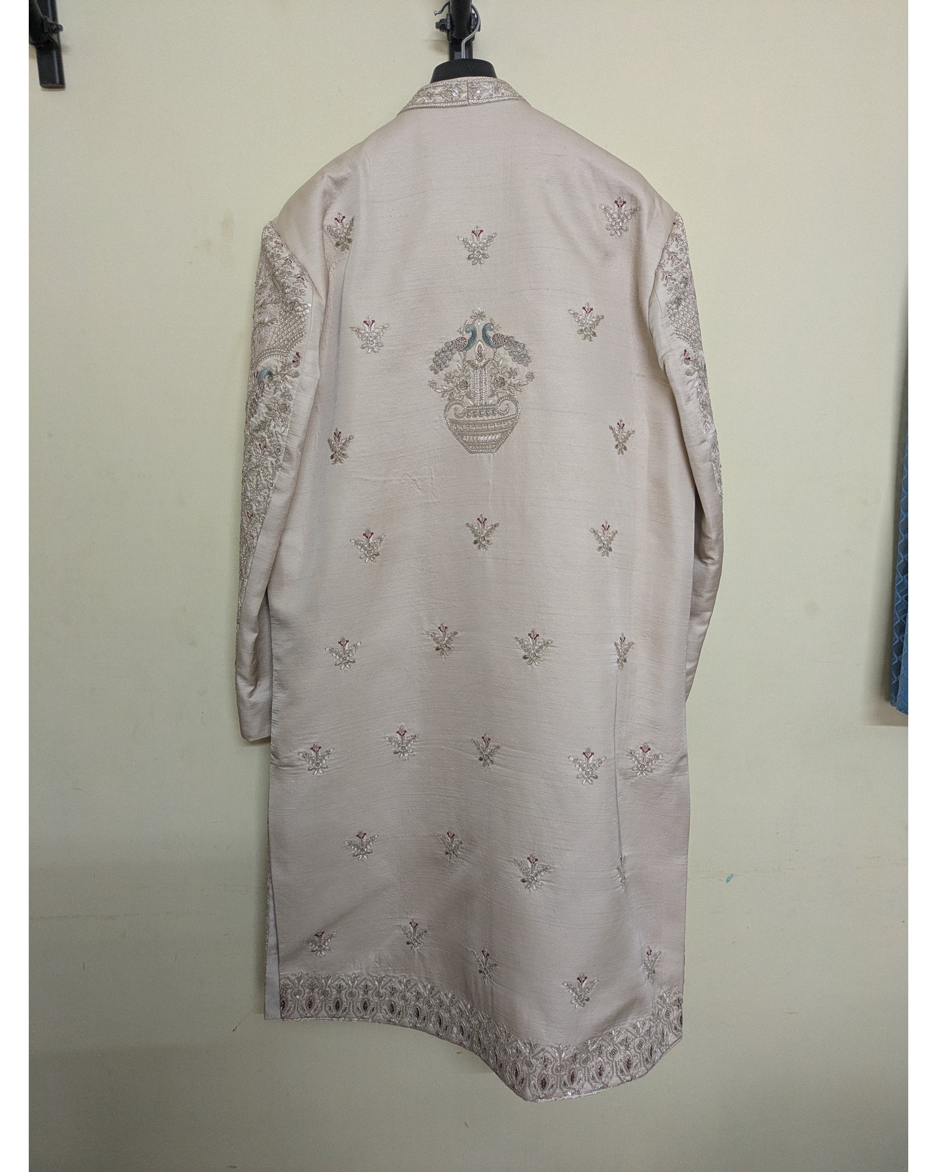 A light beige traditional long coat or kurta with intricate silver and gold embroidery, hanging on a black hanger against a plain off-white wall.