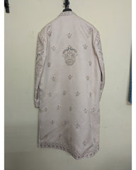 A light beige traditional long coat or kurta with intricate silver and gold embroidery, hanging on a black hanger against a plain off-white wall.