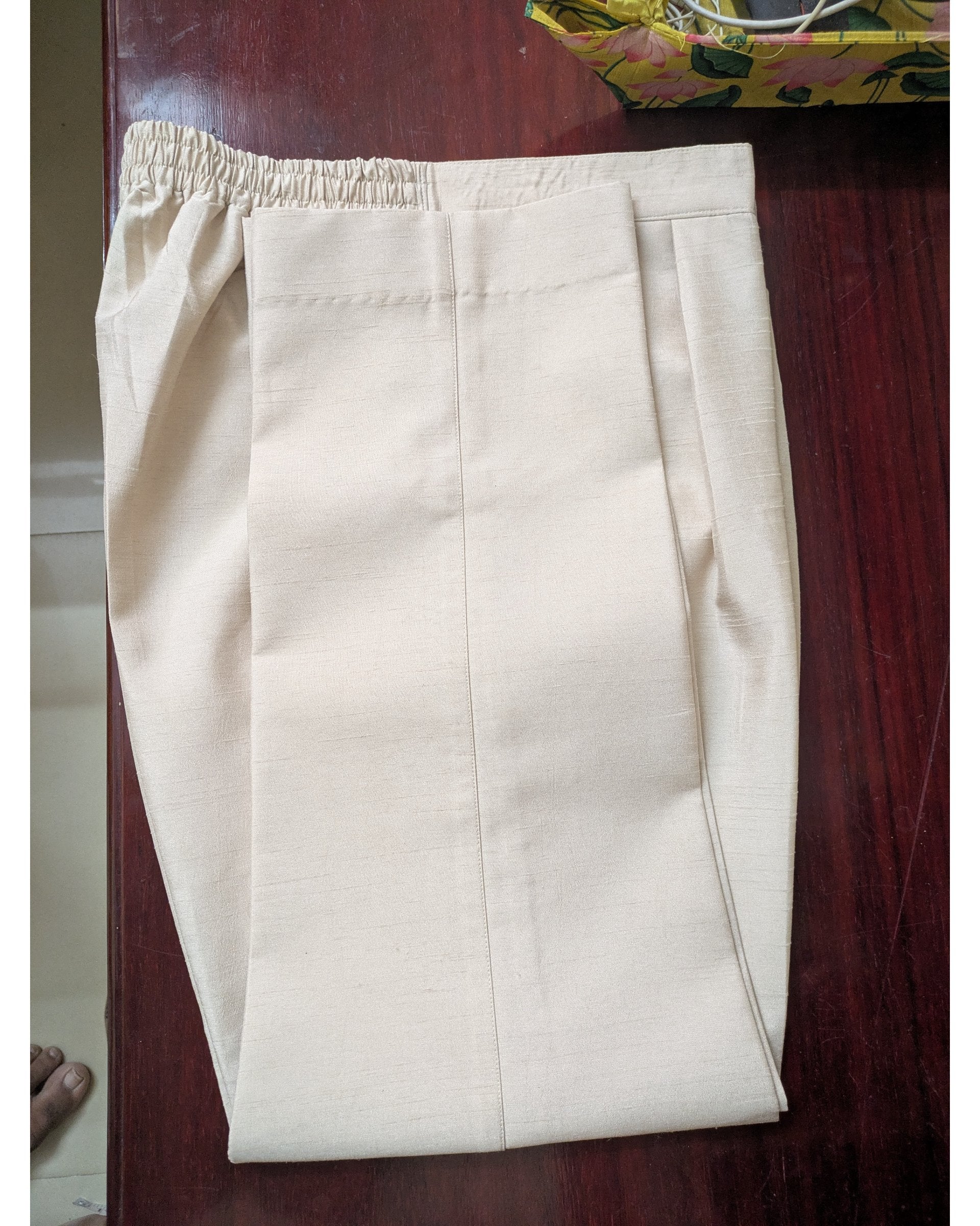 A pair of light beige trousers with an elastic waistband, folded and laid flat on a dark reddish-brown wooden surface.