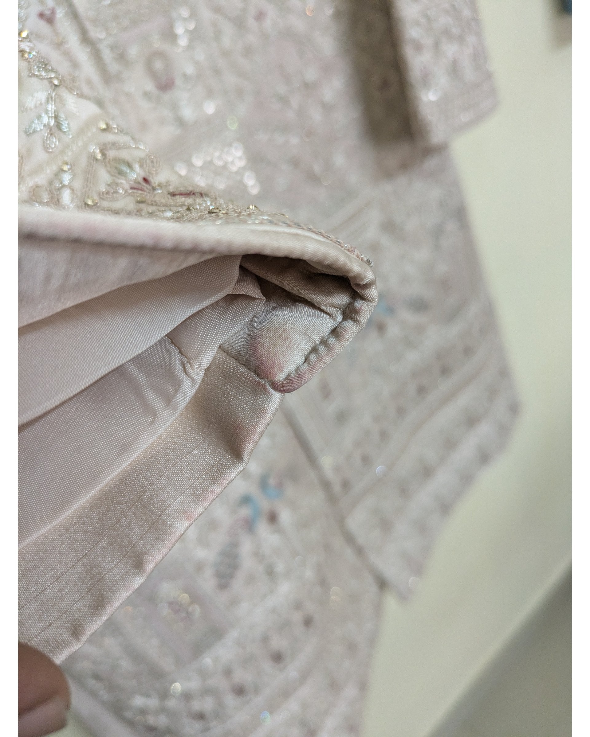 Close-up detail of a light beige embroidered fabric garment, showing a folded hem with satin-like trim and sequined embellishment. Slight stain marks.