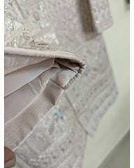 Close-up detail of a light beige embroidered fabric garment, showing a folded hem with satin-like trim and sequined embellishment. Slight stain marks.