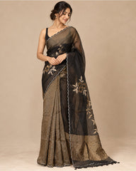 A woman modeling a black and gold saree with floral embroidery and a pleated pattern, set against a solid beige studio background.