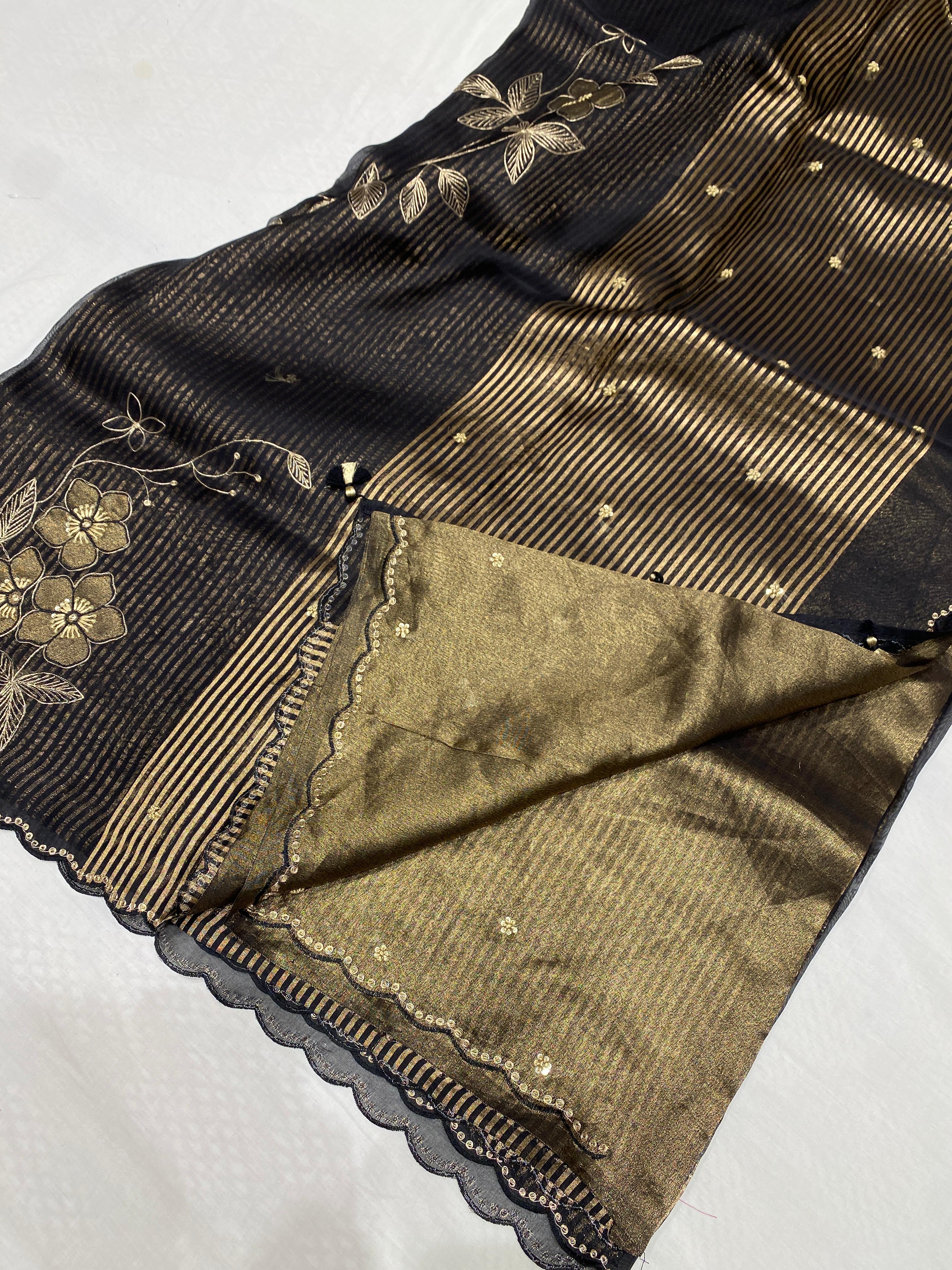 A close-up of a black and gold Banarasi silk saree, showing the black body with gold floral zari embroidery, a wide gold striped border, and the matching gold-gilt inner fabric with scalloped edges.