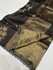 A close-up of a black and gold Banarasi silk saree, showing the black body with gold floral zari embroidery, a wide gold striped border, and the matching gold-gilt inner fabric with scalloped edges.