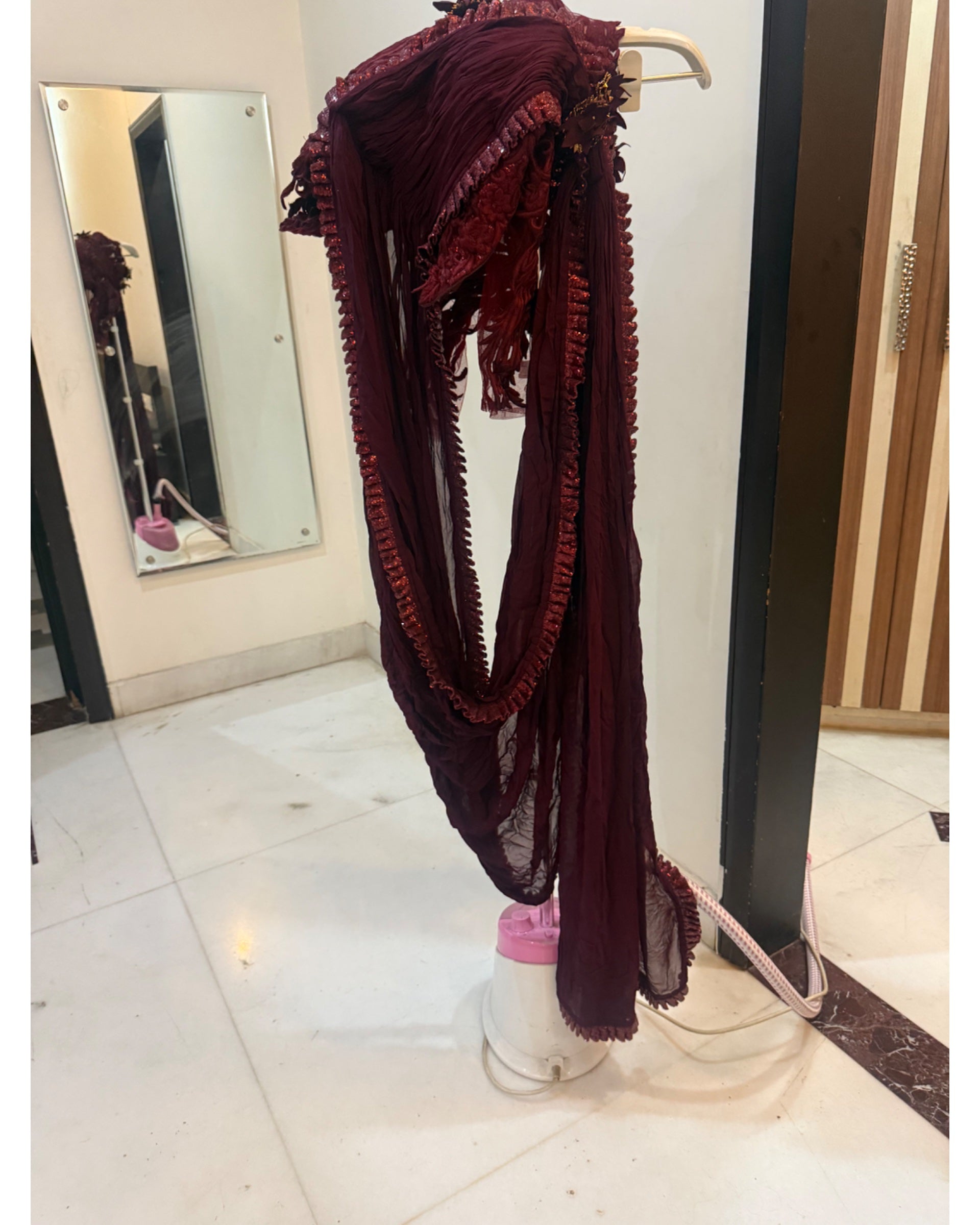 A long, sheer burgundy fabric garment or scarf with ruffled trim is draped over a white stand in an indoor room with tiled floors and a mirror.