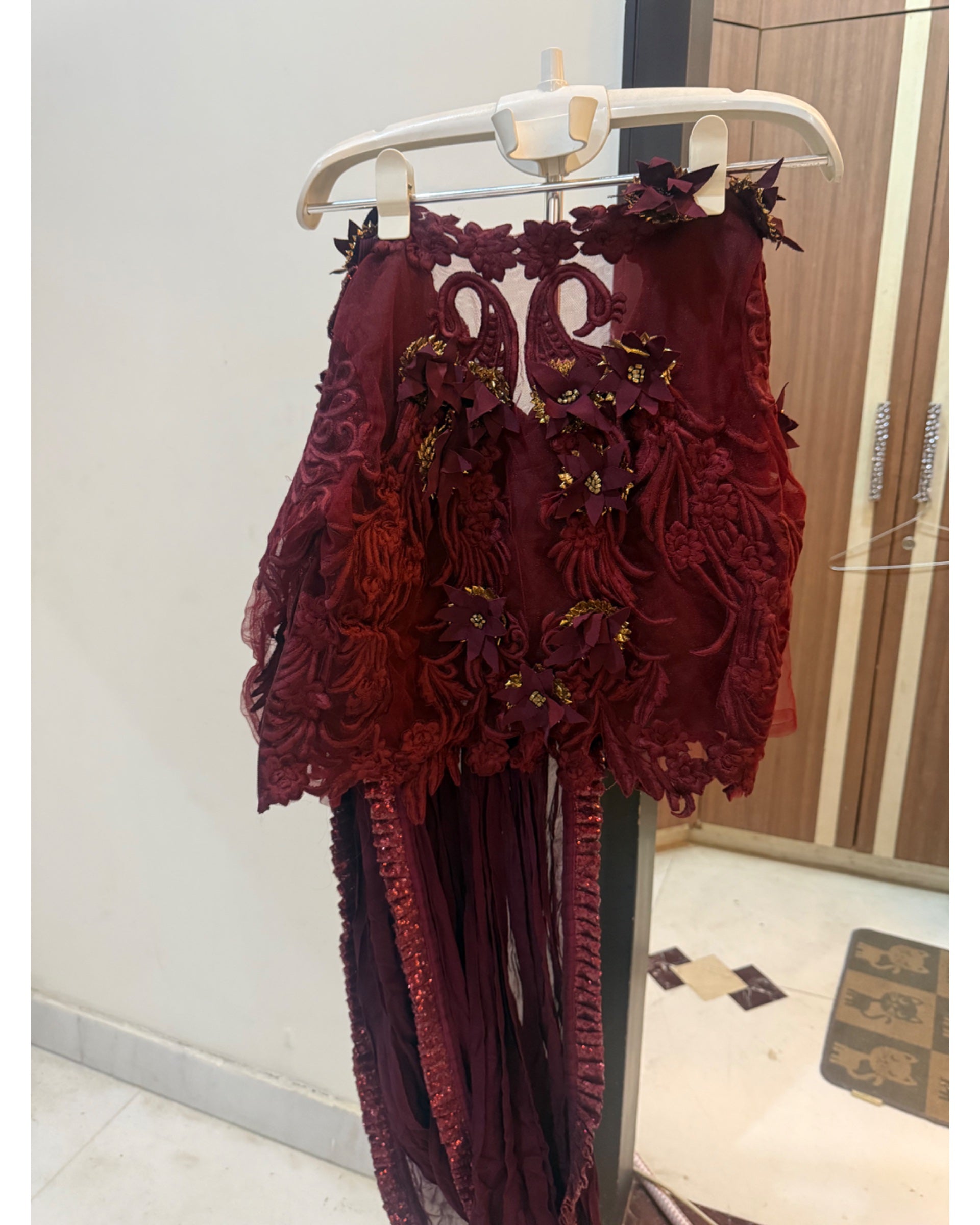 A deep burgundy lace and mesh dress with 3D floral appliques and gold embroidered details, hanging on a white plastic hanger in an indoor room.