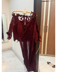 A deep burgundy red dress with intricate lace appliques and sheer draped panels, hanging on a wooden hanger in an indoor setting.