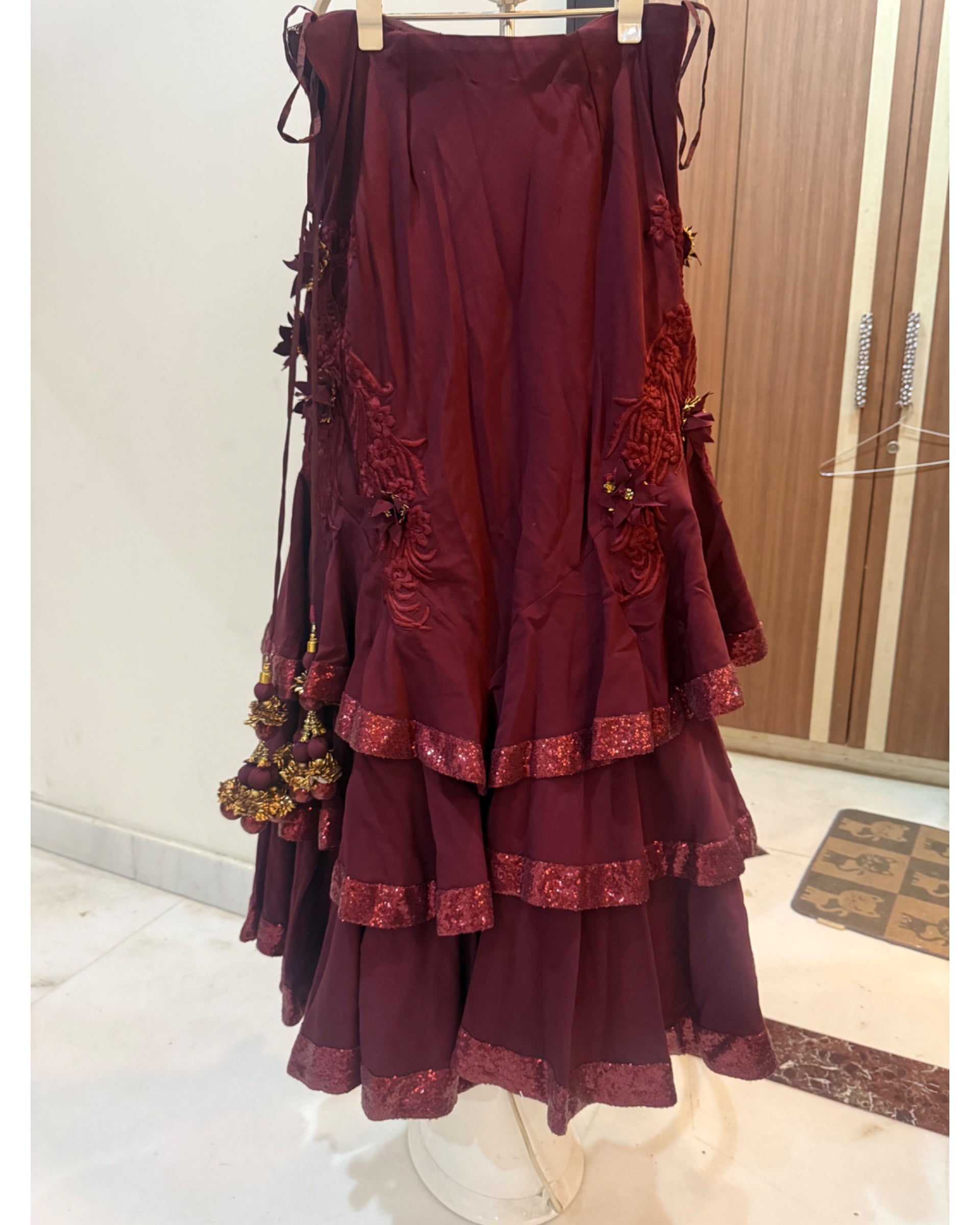A tiered lehenga in deep maroon with gold floral embroidery and matching lace trim, hanging on a white stand in an indoor room.