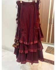 A tiered lehenga in deep maroon with gold floral embroidery and matching lace trim, hanging on a white stand in an indoor room.