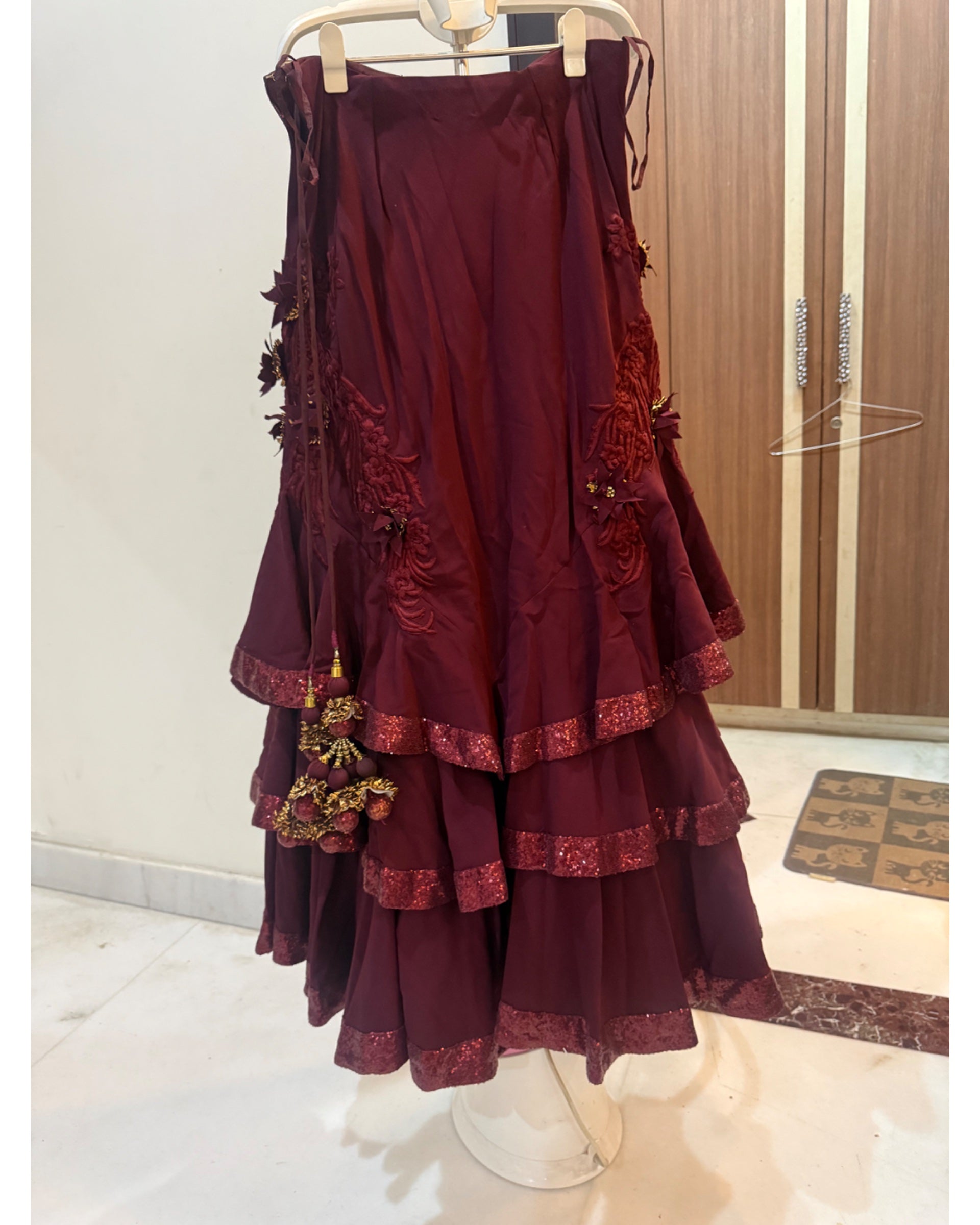 A tiered maroon lehenga skirt displayed on a hanger stand in an indoor boutique setting. The lehenga features floral embroidery on the sides and a matching embroidered tassel charm, with lace-trimmed ruffled edges.