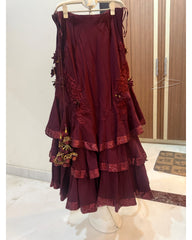 A tiered maroon lehenga skirt displayed on a hanger stand in an indoor boutique setting. The lehenga features floral embroidery on the sides and a matching embroidered tassel charm, with lace-trimmed ruffled edges.