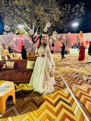 Woman in a white and gold traditional outfit standing in an outdoor decorated area with people and decor elements.