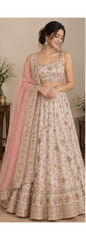 A woman models a traditional Indian lehenga choli set in a warm beige and dusty pink color palette, featuring intricate gold embroidery, a floral print, and a matching pink dupatta.
