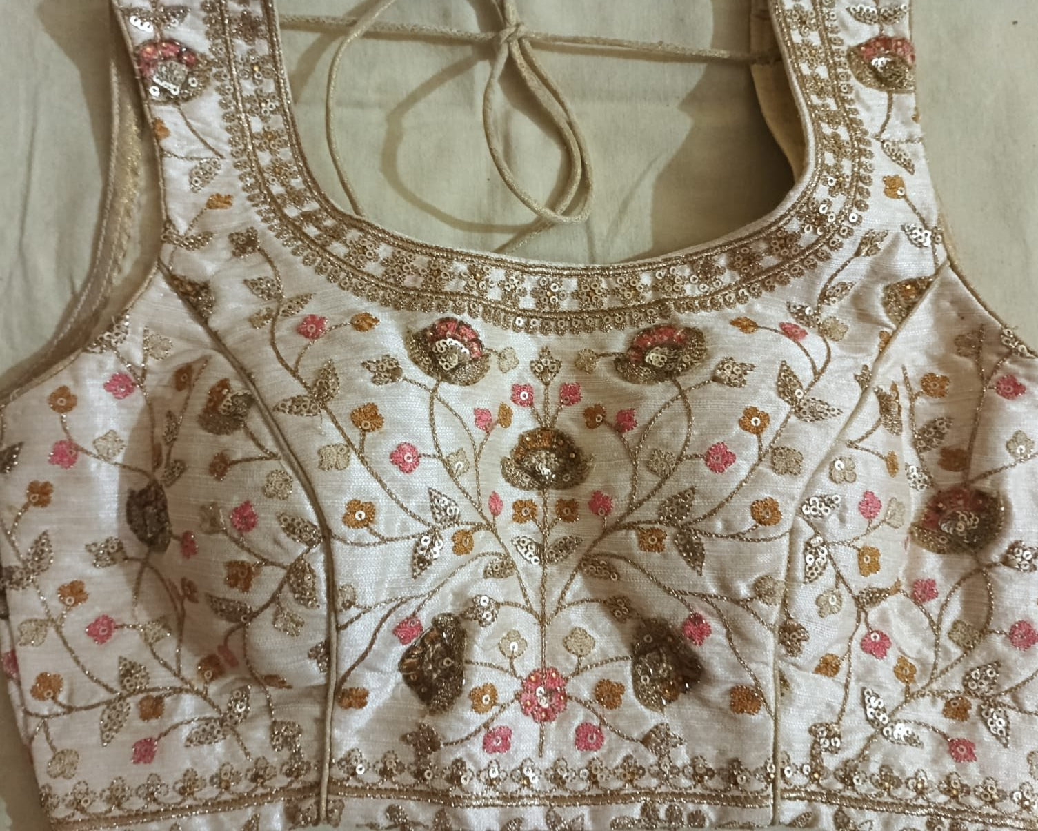 Close-up of a cream-colored embroidered blouse or sari fabric with metallic gold floral embroidery, pink floral accents, and sequined details, laid flat on a neutral fabric background.