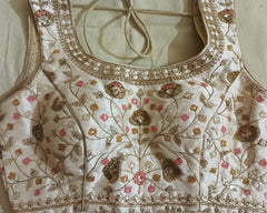 Close-up of a cream-colored embroidered blouse or sari fabric with metallic gold floral embroidery, pink floral accents, and sequined details, laid flat on a neutral fabric background.
