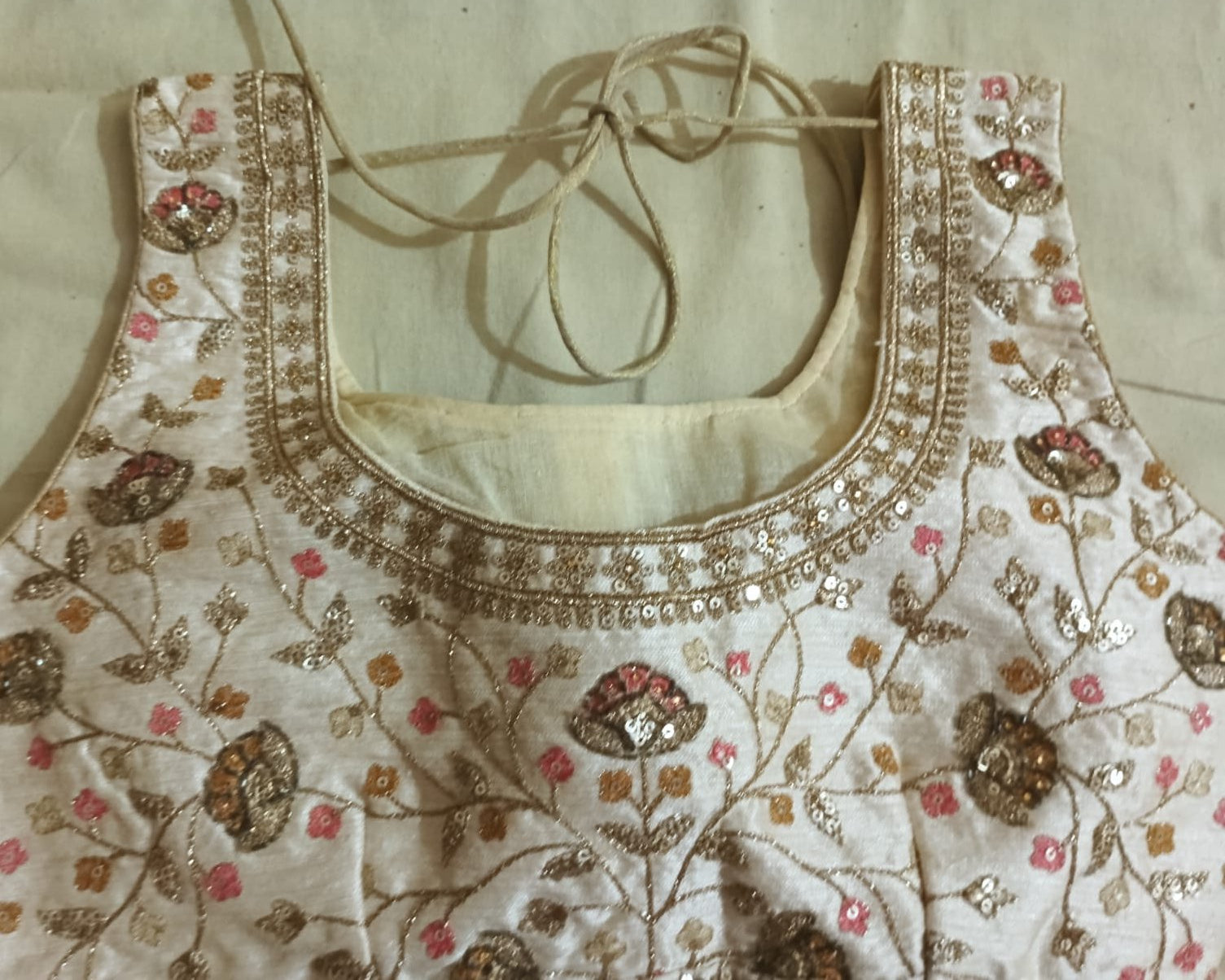 An embroidered blouse bodice laid flat on a light beige fabric background. The off-white fabric features intricate gold metallic embroidery, sequins, and floral motifs in pink, gold, and brown, with a tie-neck design.