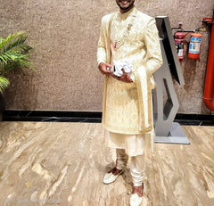 Man in a traditional cream kurta standing indoors with a plant and fire extinguishers in the background.