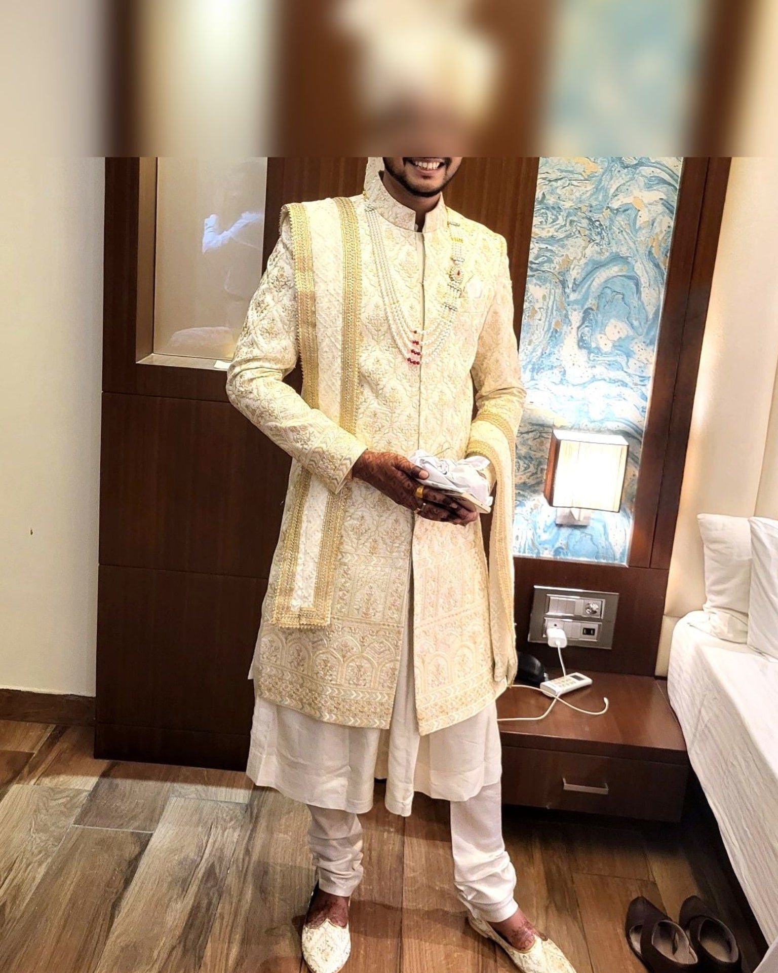 Man in traditional white sherwani standing in a room with wooden floor and furniture.