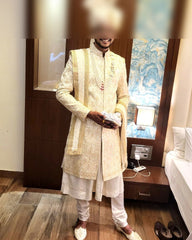 Man in traditional white sherwani standing in a room with wooden floor and furniture.