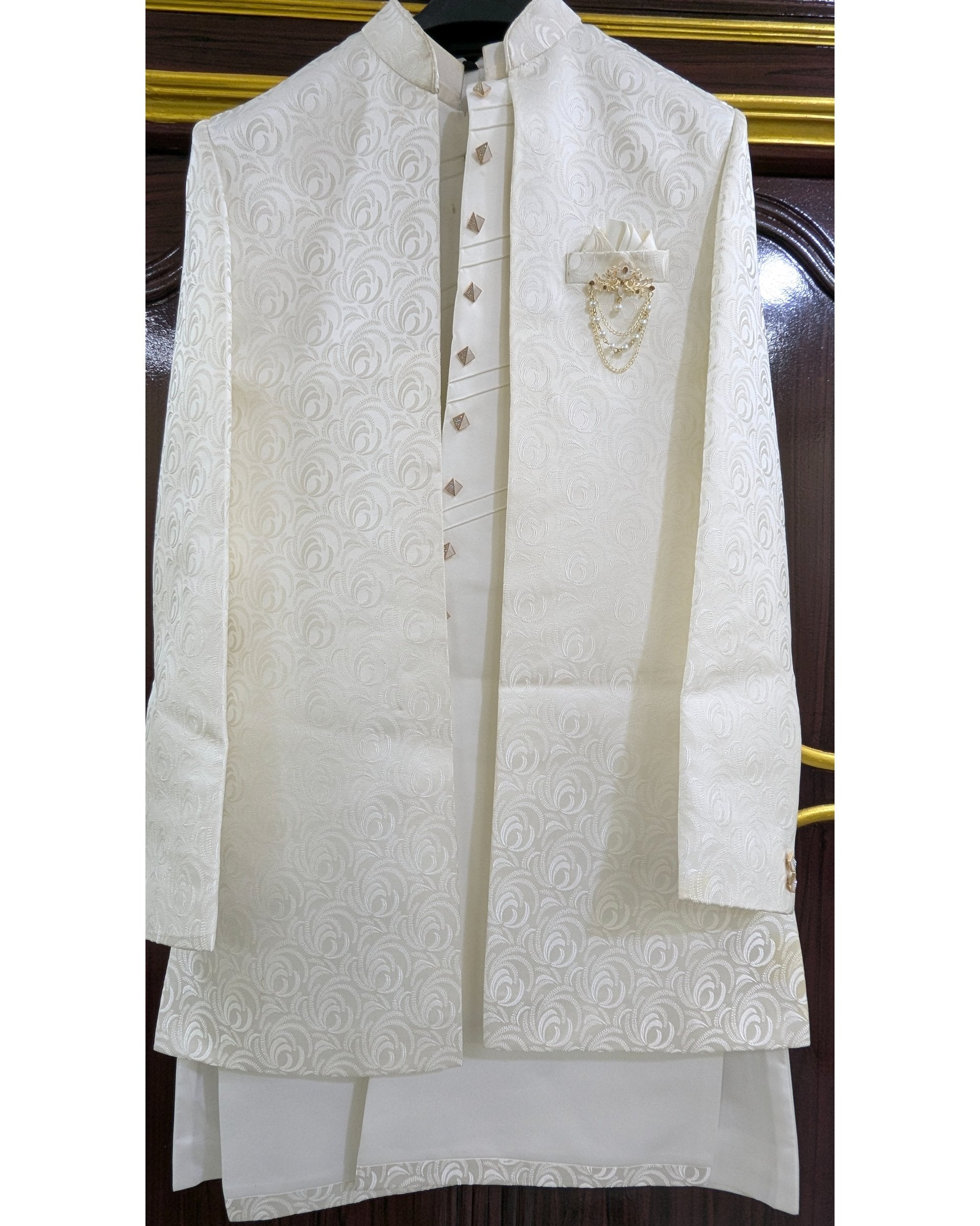 An off-white/cream traditional men's outfit consisting of a long-sleeved embroidered jacket and a matching kurta, displayed on a hanger against a dark wooden door background.