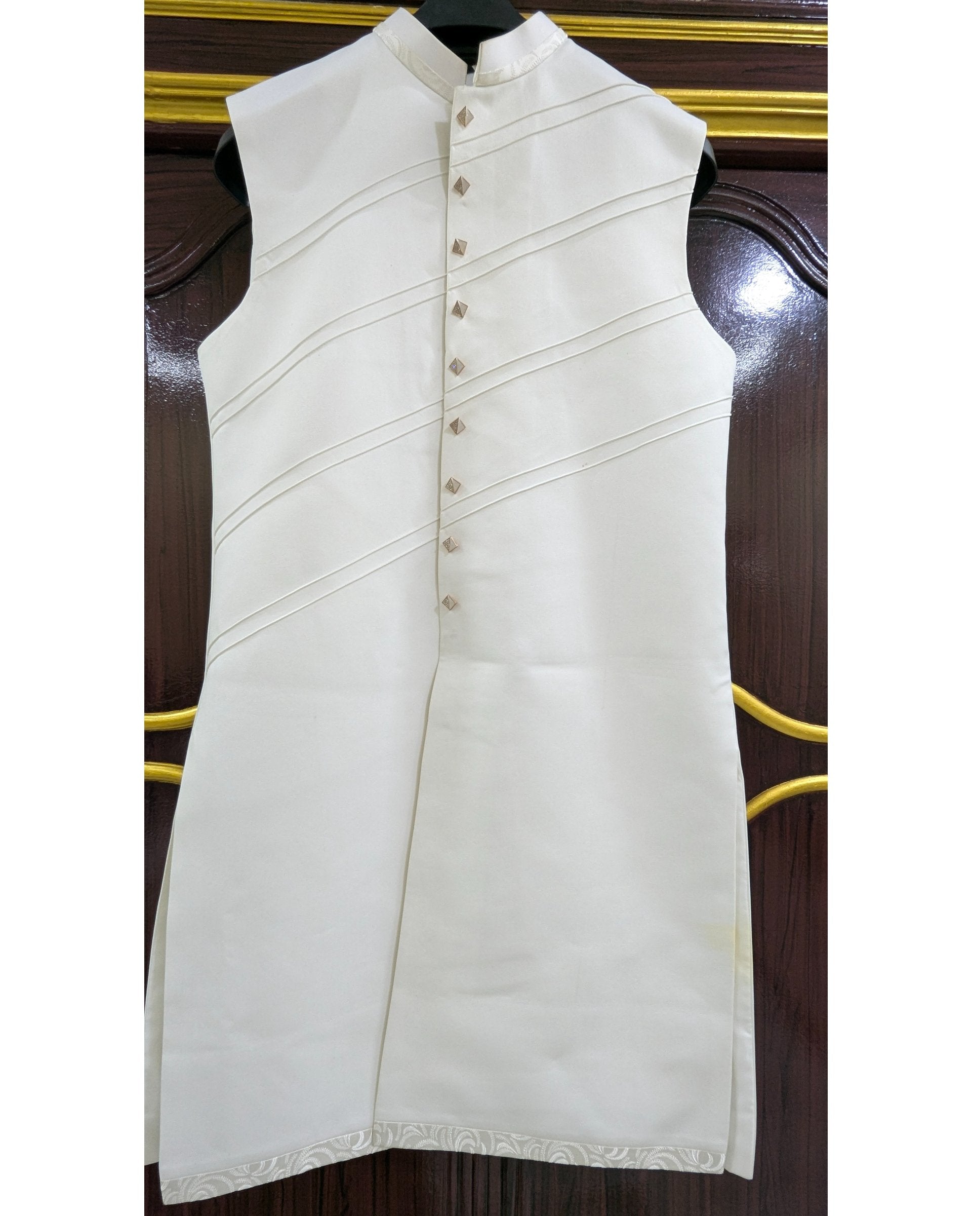 An off-white sleeveless kurta with a mandarin collar and decorative diagonal pleats, displayed on a black hanger against a dark wood background.