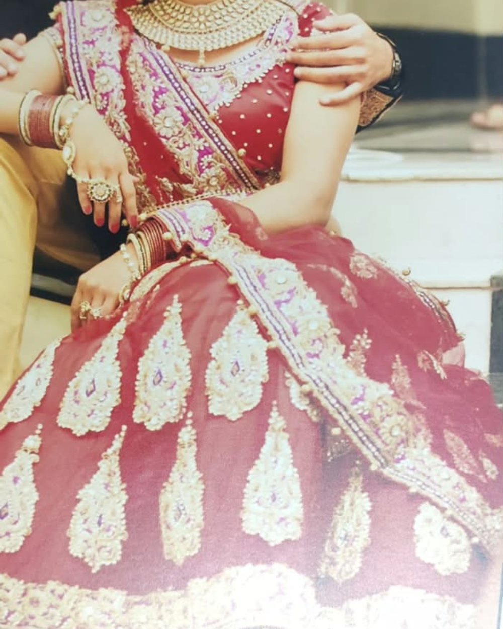 A close-up of a person wearing an ornate traditional red lehenga choli outfit with heavy gold and silver embroidery, embellished with teardrop motifs and draped over the shoulder.
