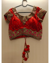 A red traditional blouse with intricate gold and silver embroidery, hanging on a black hanger against a plain beige wall.