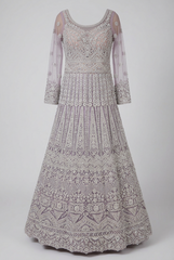 A heavily embellished floor-length gown with intricate beadwork and embroidery in pale lavender and silver tones, featuring sheer long sleeves and a flared skirt, shown against a light gray studio background.