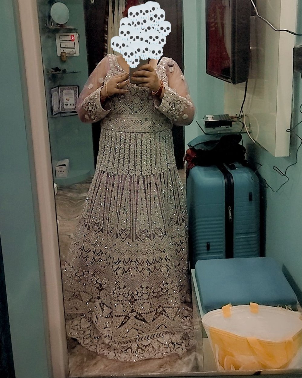 A woman takes a mirror selfie wearing a long, heavily embellished lace dress or gown in a beige/taupe tone. The photo is taken in a room with a blue suitcase, a yellow bag, and a television visible in the background.