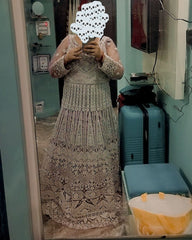 A woman takes a mirror selfie wearing a long, heavily embellished lace dress or gown in a beige/taupe tone. The photo is taken in a room with a blue suitcase, a yellow bag, and a television visible in the background.
