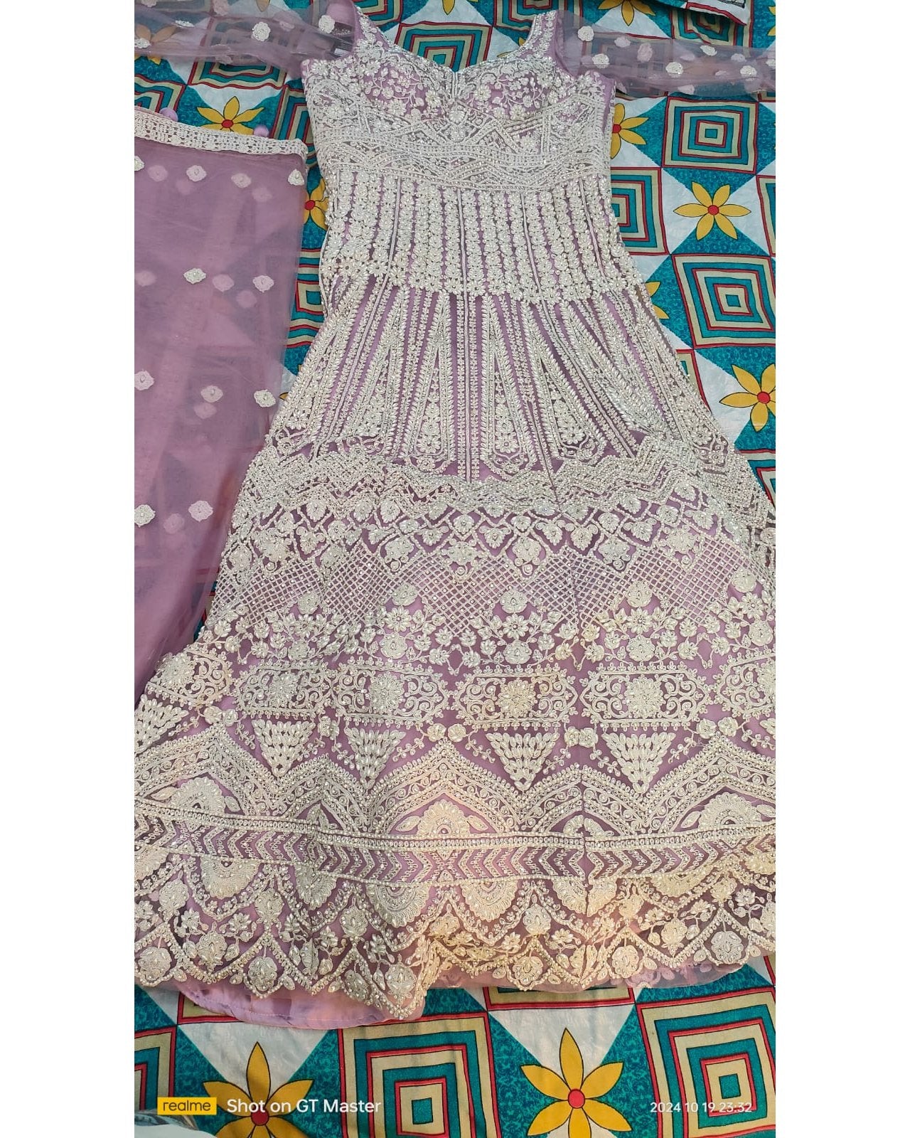 A lavender and off-white heavily embroidered sleeveless dress or anarkali-style gown laid flat, with a matching sheer embroidered dupatta partially visible on the left. The garment features intricate floral and geometric lace patterns across the bodice and full skirt.