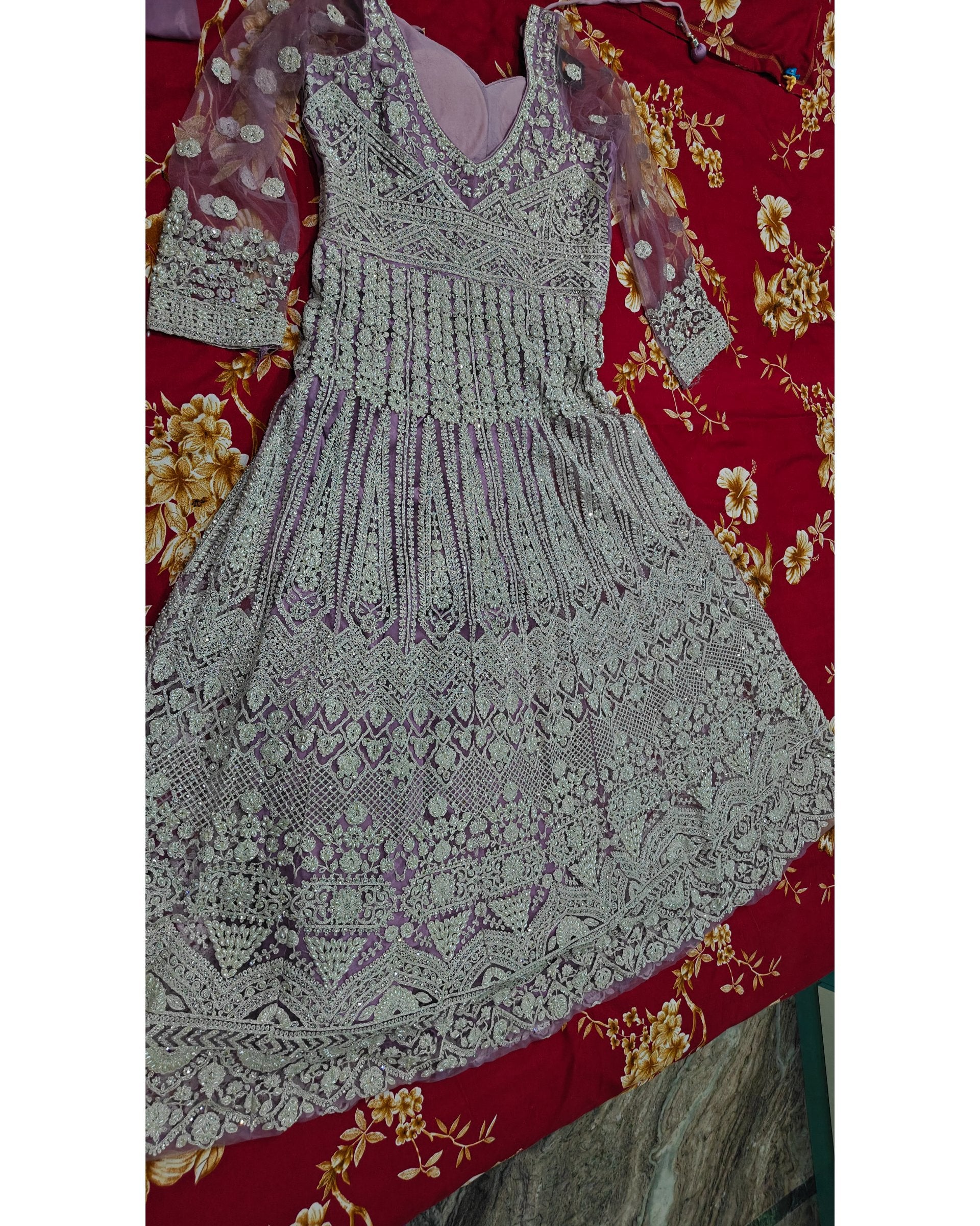 A heavily embellished lavender and silver embroidered dress laid flat on a red floral fabric background.