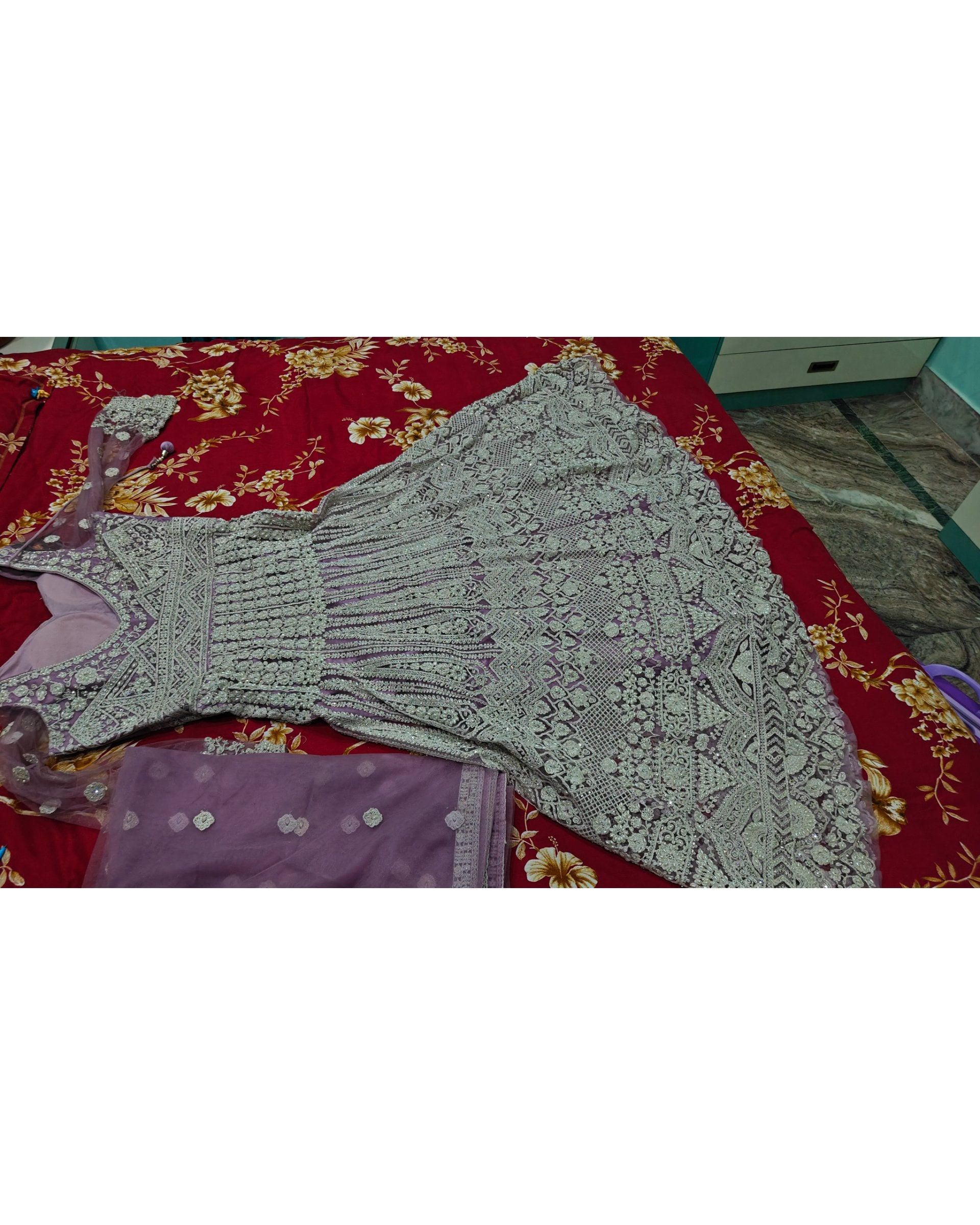 A light gray embroidered dress with a matching purple dupatta and folded fabric pieces, laid out on a red floral bedsheet.