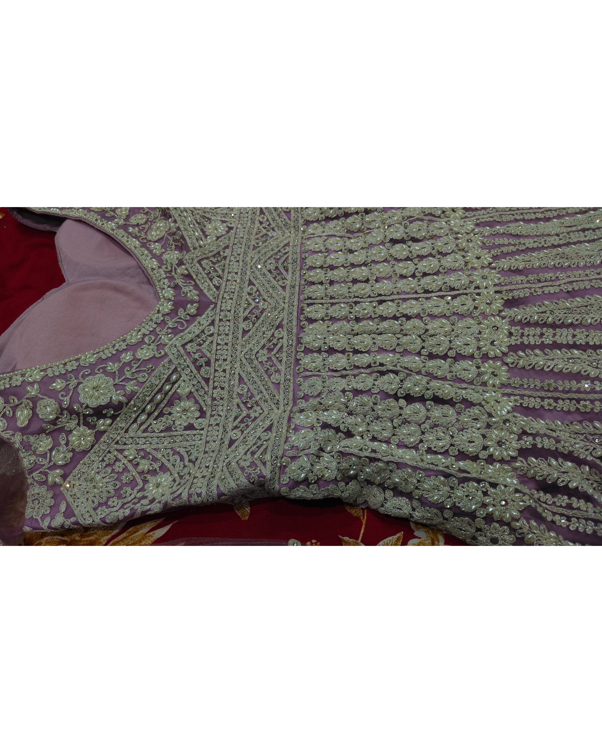 A close-up of a heavily embroidered traditional garment, showing intricate light green sequin and bead work on a dusty purple fabric, laid flat on a red surface.