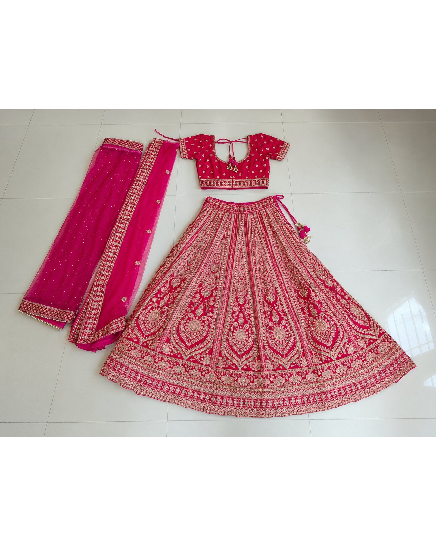 A pink and red embroidered Indian ethnic wear set consisting of a flared lehenga skirt, a short-sleeve blouse, and a dupatta, laid flat on a light-colored tiled floor.