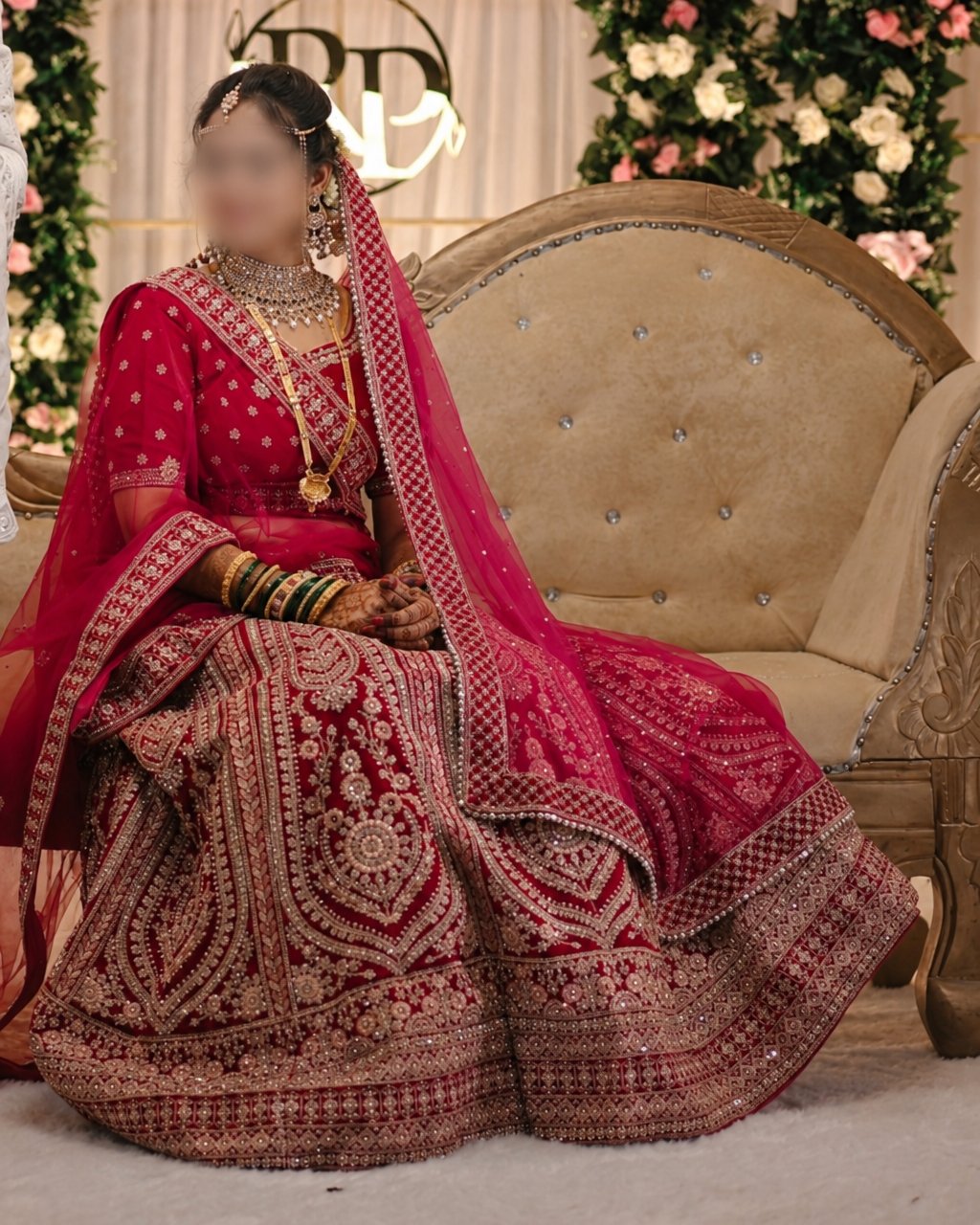 A woman seated on a tufted armchair wearing an ornate red and pink embroidered traditional outfit with a matching sheer drape, gold jewelry, and a heavily embellished skirt. The setting includes floral decor and a cream backdrop.